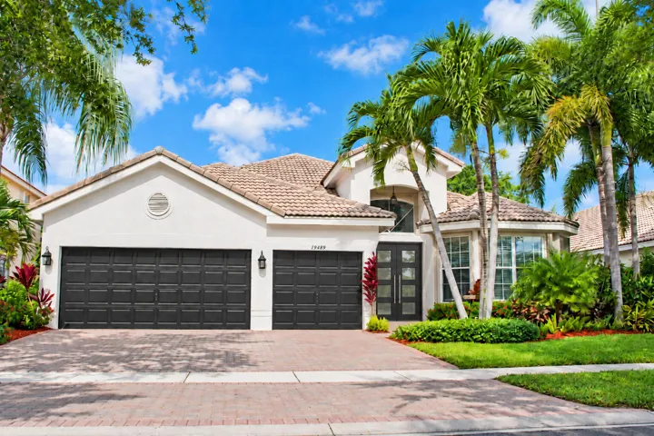 Stucco exterior with a tile roof, brick paver driveway, two dark garage doors, and mature tropical landscaping