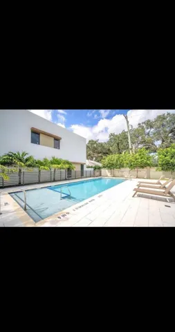 Rectangular swimming pool with light-colored decking, surrounded by a low fence and lush landscaping, featuring a modern building in the background
