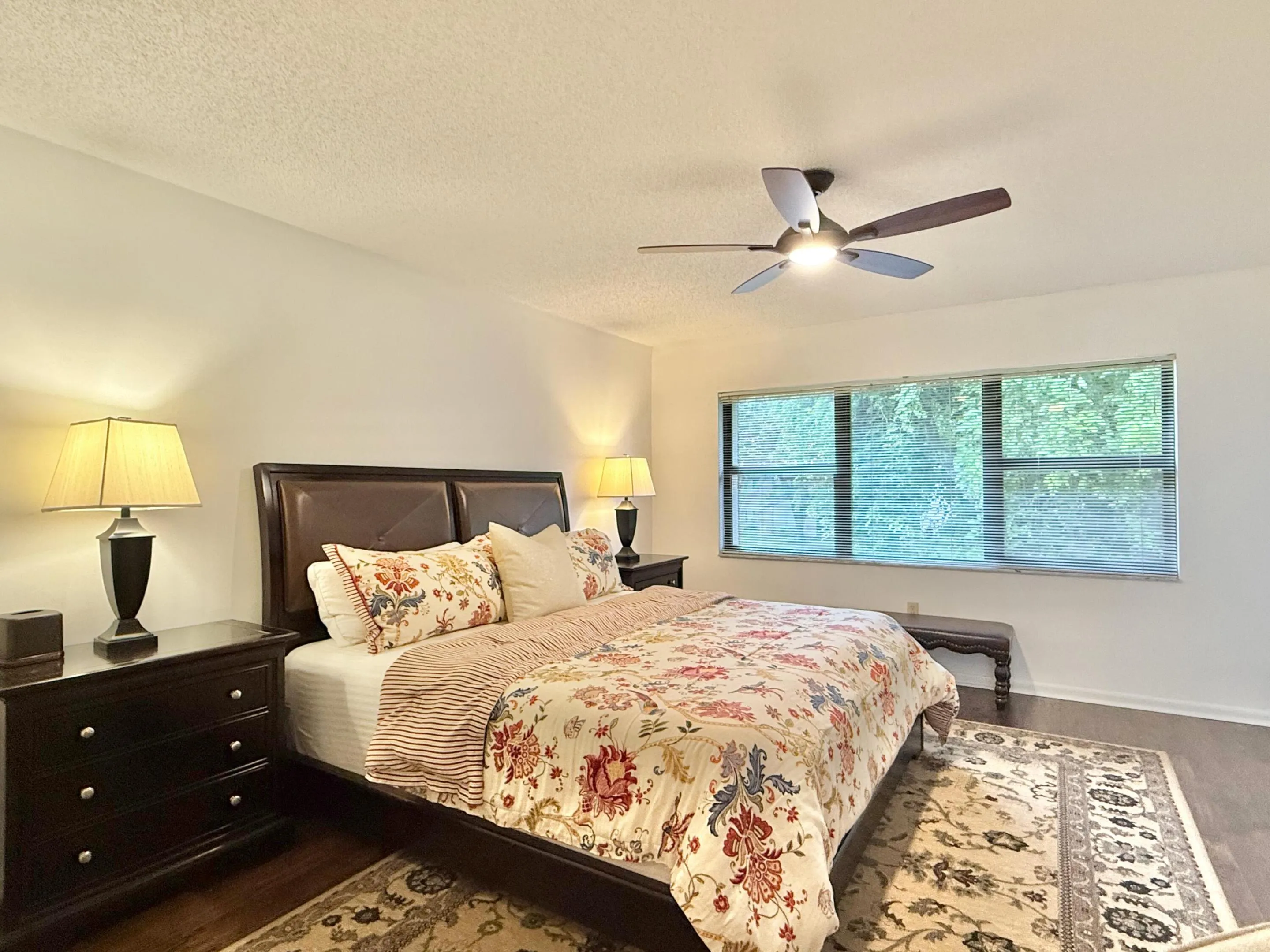 Wood-finish flooring, broad window with horizontal blinds, and a ceiling fan with integrated lighting