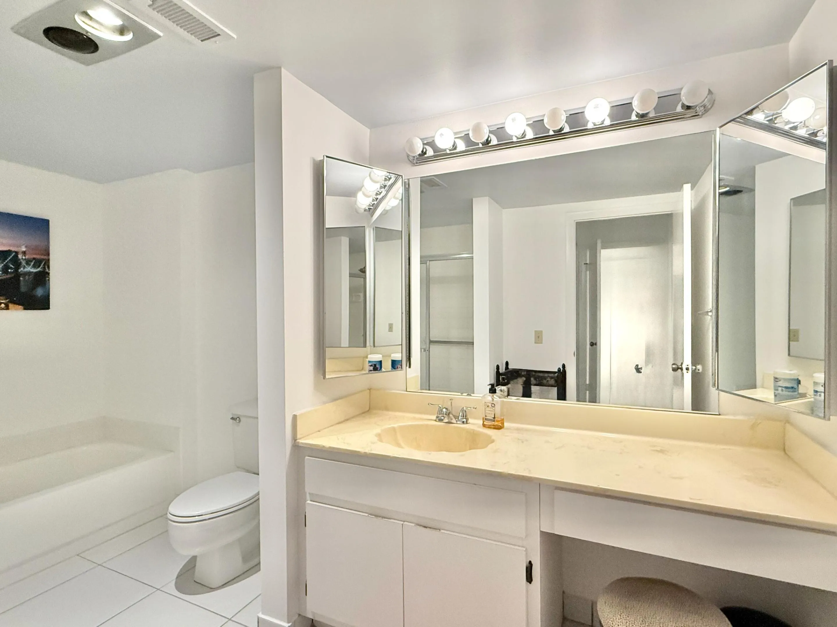 Vanity with an integrated sink and light-toned countertop, large wall-mounted mirror with integrated lighting, white tiled flooring, and a built-in bathtub