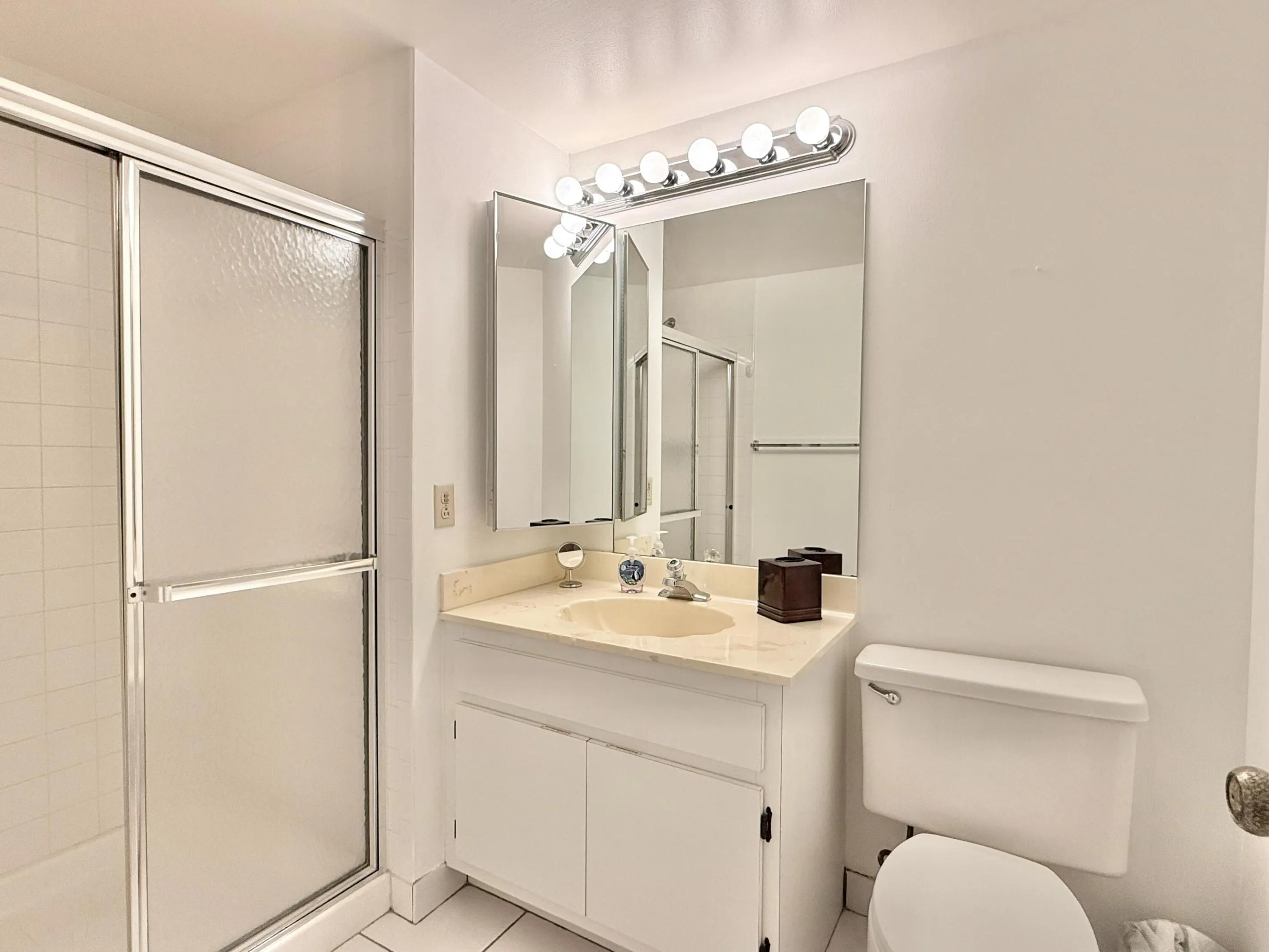 White vanity with an integrated sink and light-toned countertop, framed mirror with vanity lighting, and a shower with sliding frosted glass doors