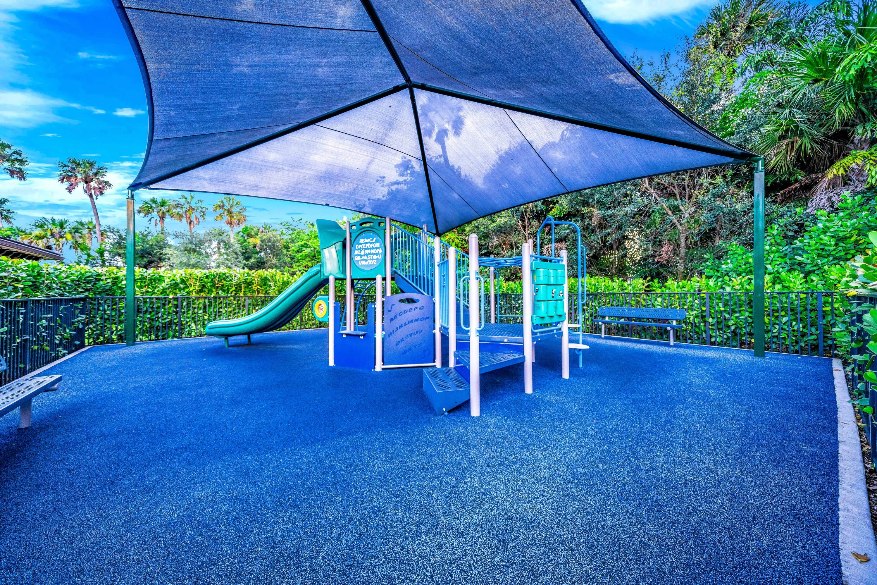 Recreational area featuring a shade structure, play equipment with slides, a blue cushioned ground surface, and perimeter fencing