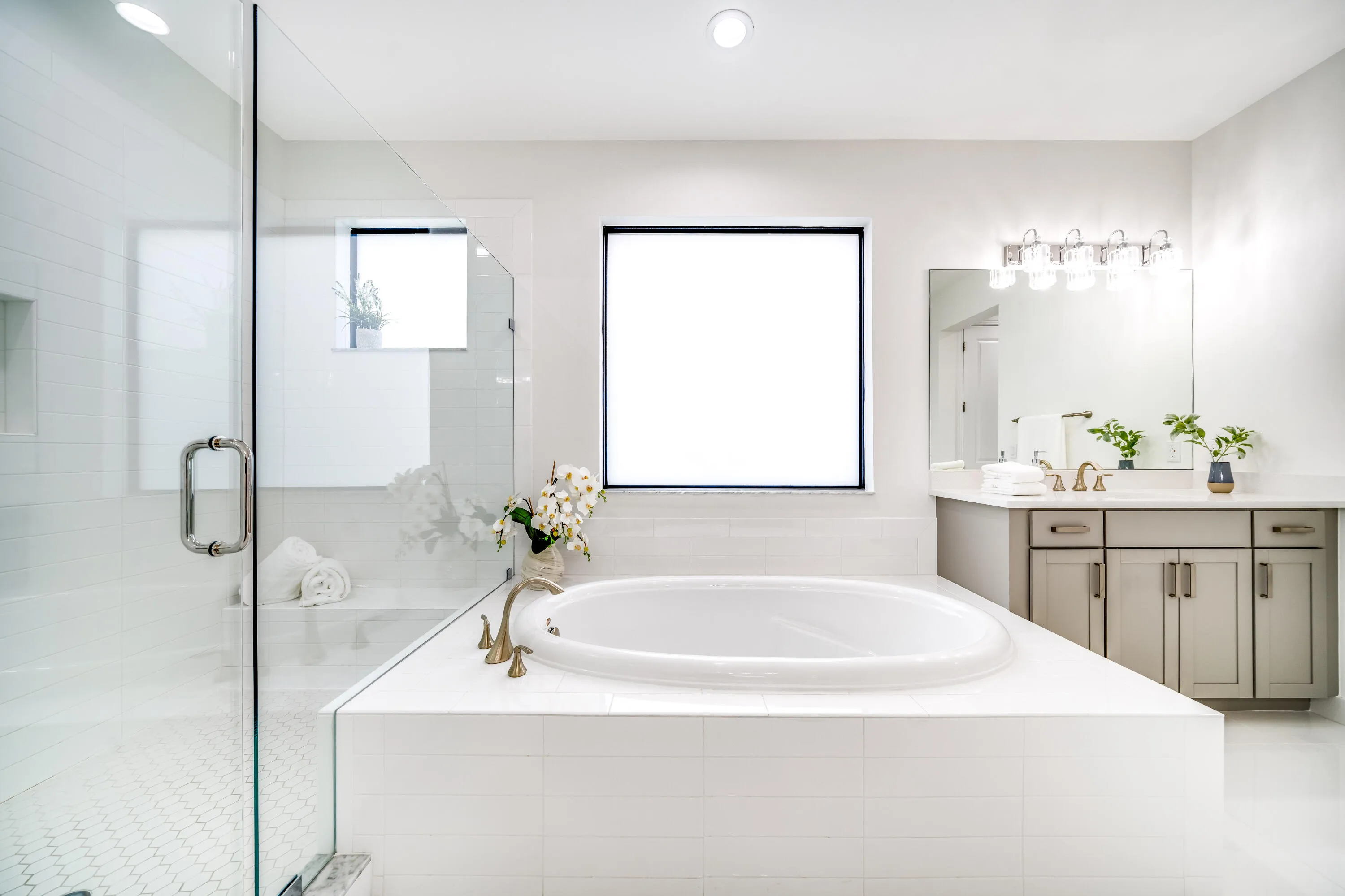 Freestanding soaking tub with brass-finish fixtures, dual sink vanity with stone countertop, glass-enclosed shower with tiled walls, and large windows