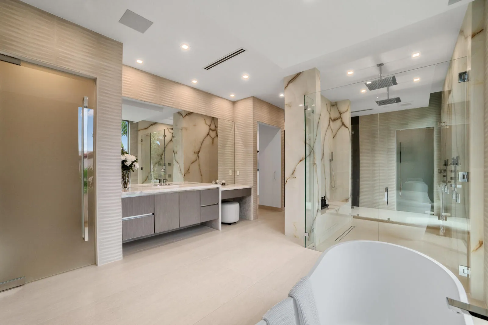 Luxurious primary bath showcasing an oversized glass-enclosed shower with dual rain heads, book-matched stone, floating vanities, and a dedicated makeup station.