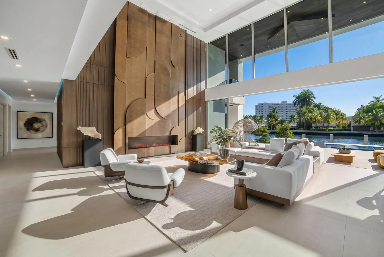 Soaring two-story great room framed by floor-to-ceiling glass, a dramatic paneled feature wall, linear fireplace, and captivating waterfront views.