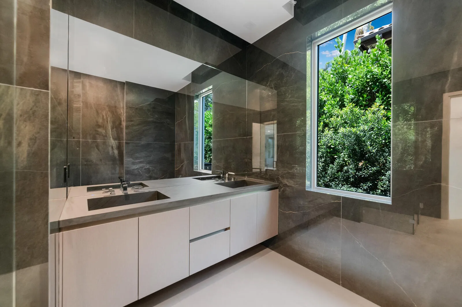 Luxurious en-suite bath finished with floor-to-ceiling stone, a floating dual-sink vanity, an oversized mirror, frameless glass shower, and abundant natural light.