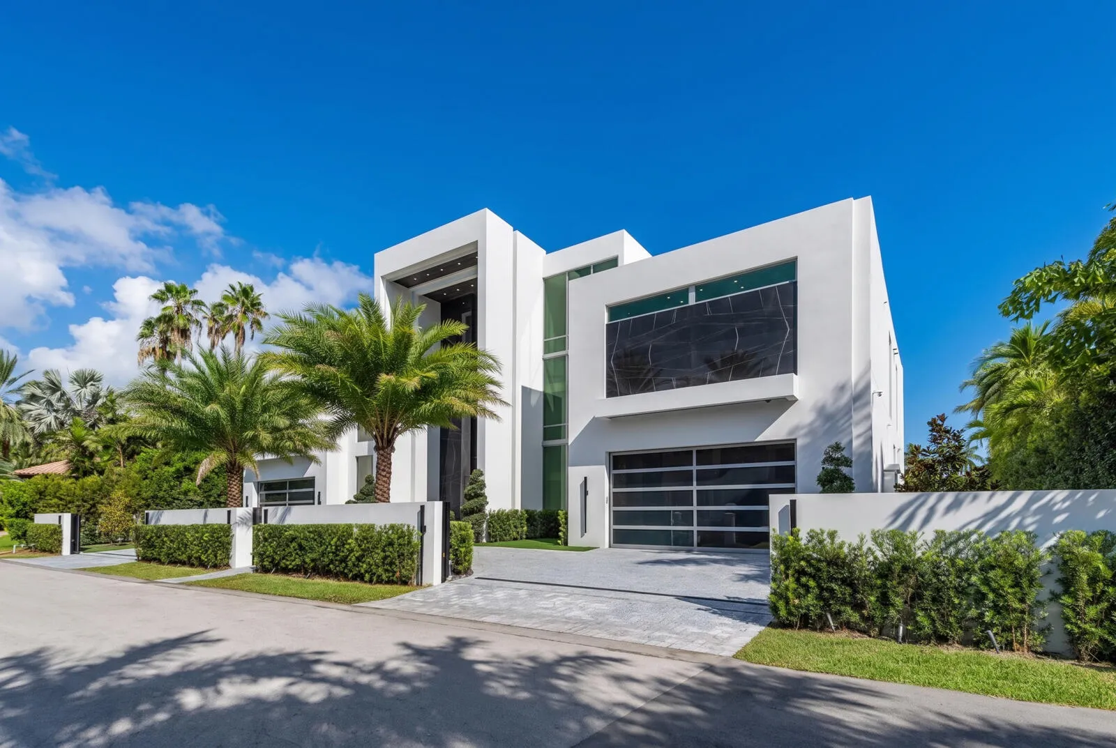Right-side exterior view showcasing bold contemporary architecture, dramatic floor-to-ceiling glass, a sleek garage, private motor court, and lush tropical landscaping.
