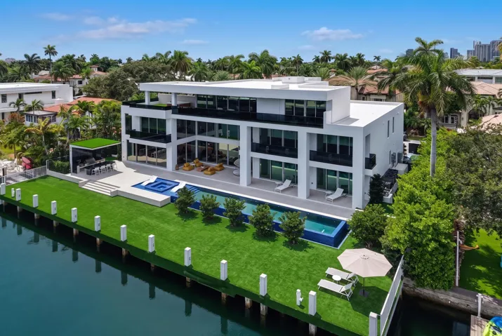 Spectacular waterfront setting featuring a 75-foot infinity-edge pool with integrated spa and submerged sun shelf, a covered summer kitchen and barbecue area, expansive outdoor living spaces, and a turf-finished concrete dock extending approximately 20 feet beyond the seawall.