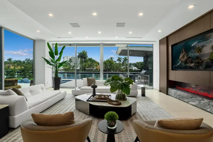 Waterfront family and TV room with custom millwork, a built-in television, linear fireplace with dramatic natural-stone surround, and floor-to-ceiling glass opening to the resort-style outdoor living area.