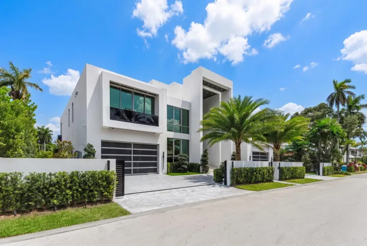 Left-side exterior view showcasing the home’s bold modern architecture, expansive floor-to-ceiling glass, sleek garage, gated motor court, and lush tropical landscaping.