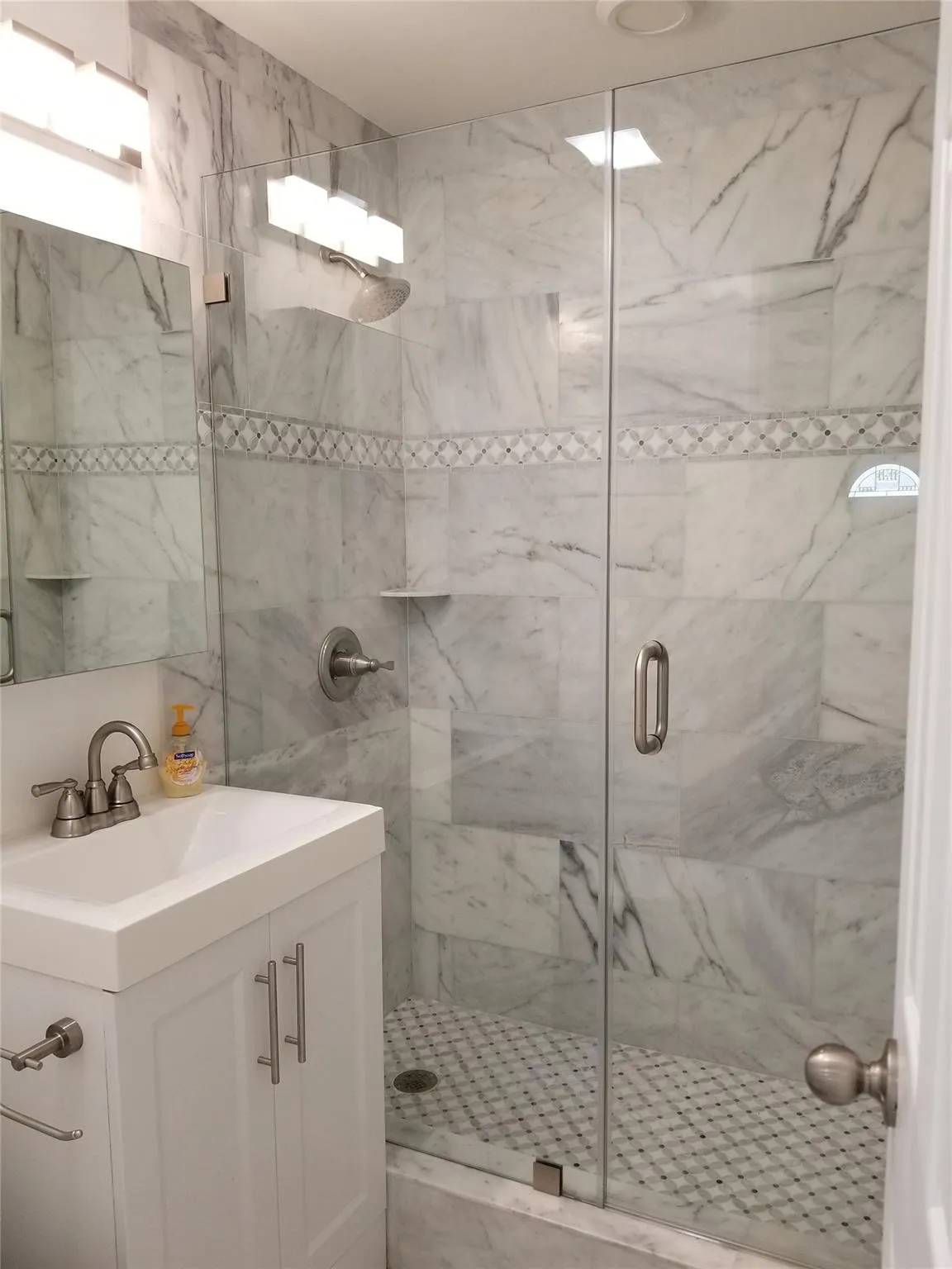 White shaker-style vanity with an integrated sink, brushed nickel faucet, frameless glass shower enclosure, shower walls featuring large-format tile and a decorative mosaic band, and a mosaic tile shower floor