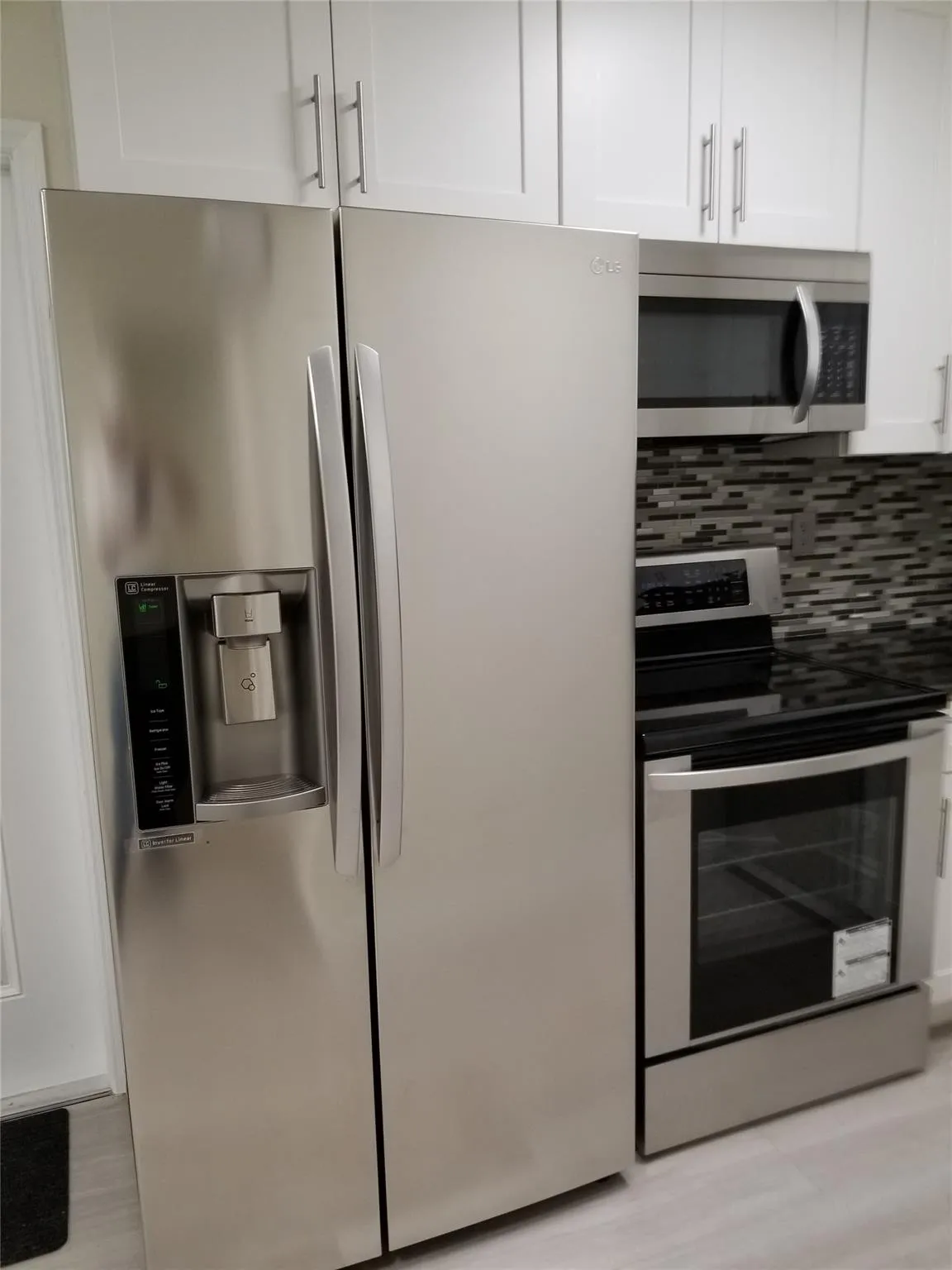 Stainless steel side-by-side refrigerator, stainless steel range with ceramic cooktop, stainless steel microwave, white upper cabinetry with bar pull hardware, and mosaic tile backsplash