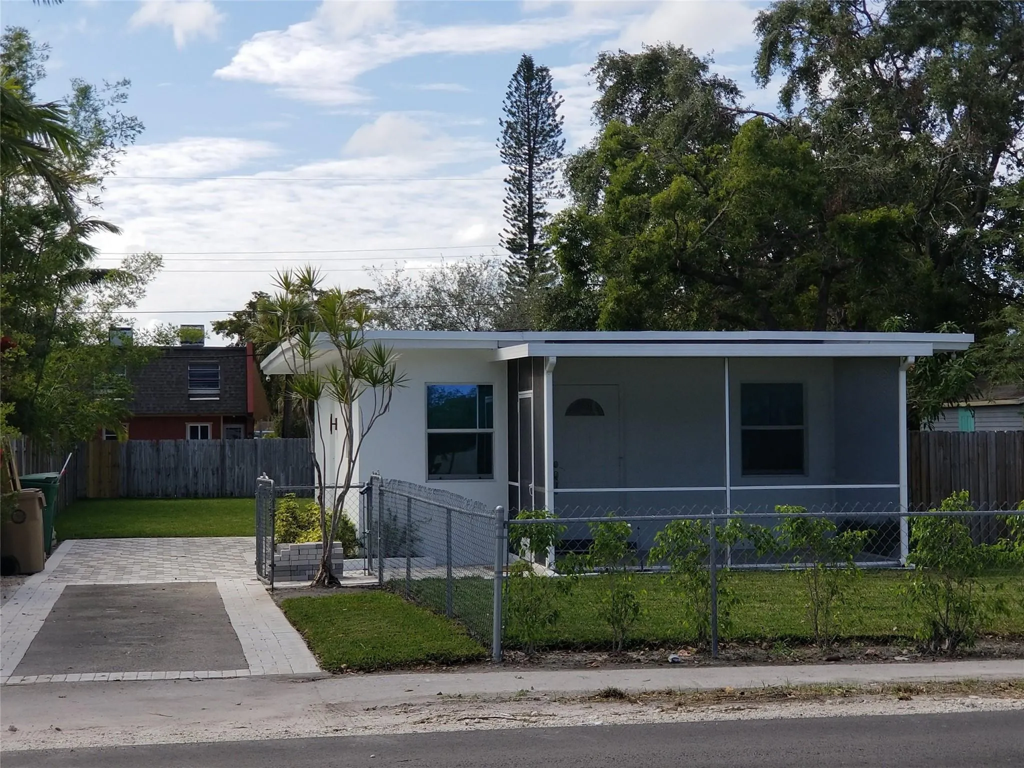 Screened front porch, white exterior, paved driveway, mature landscaping, and chain-link fence