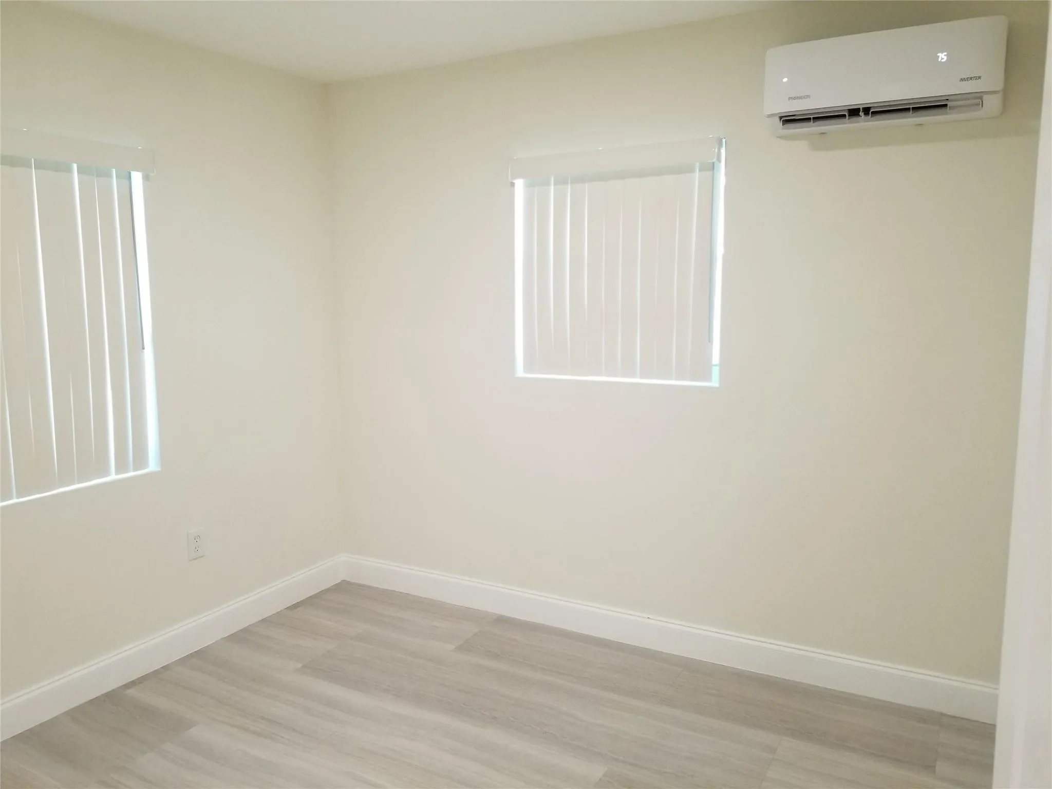 Light-toned wood-finish flooring, two windows with vertical blinds, and a wall-mounted air conditioning unit