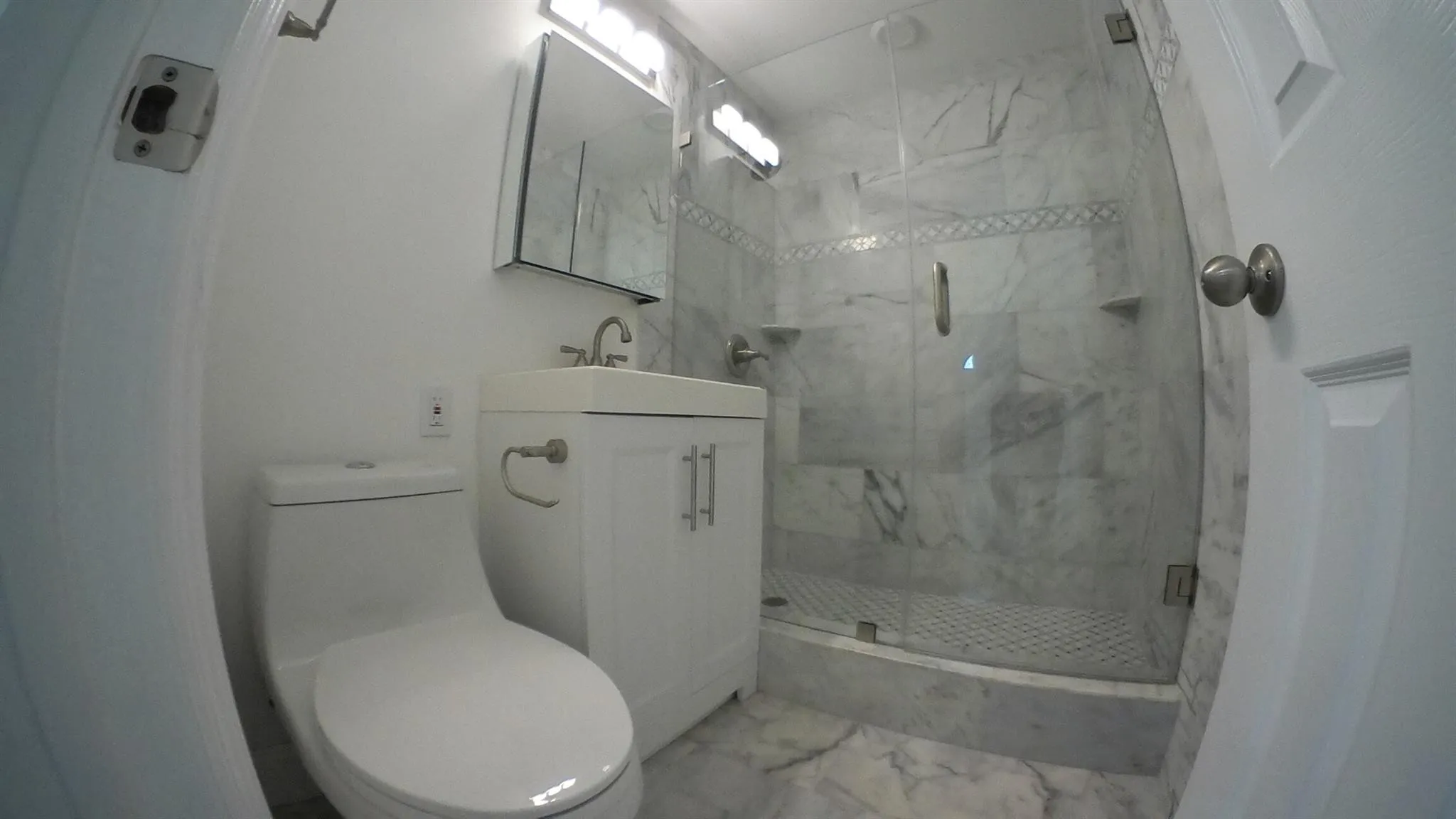 White vanity with a chrome faucet, frameless mirrored medicine cabinet, glass-enclosed shower, and full-height marble-style tile