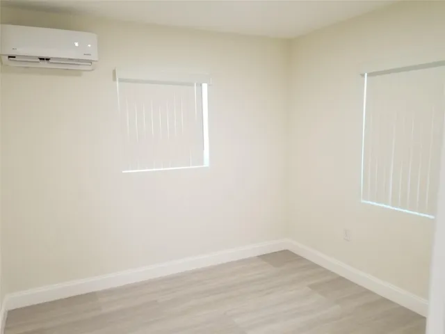 Window-mounted climate control unit, two windows with blinds, light-toned walls, and wood-finish flooring