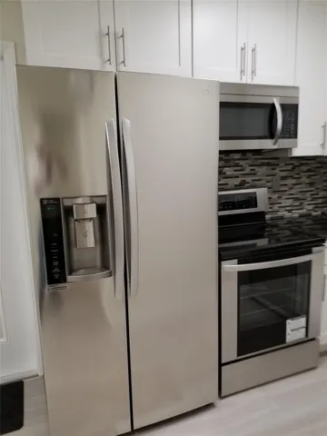 Stainless steel side-by-side refrigerator, stainless steel range with ceramic cooktop, stainless steel microwave, white upper cabinetry with bar pull hardware, and mosaic tile backsplash