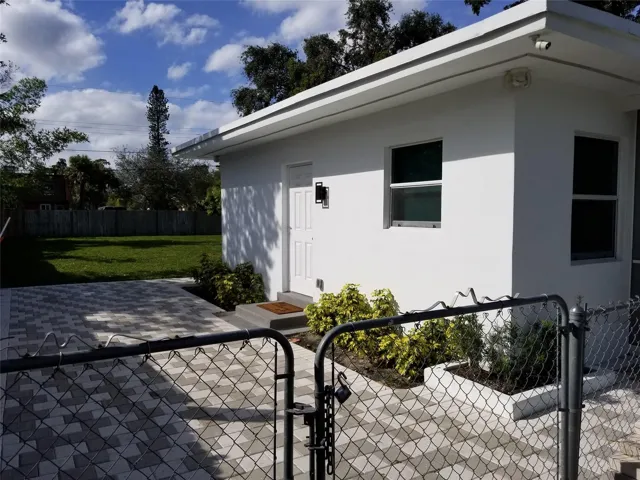 Stucco exterior with multiple windows, concrete patio with paver accents, and a chain-link fence