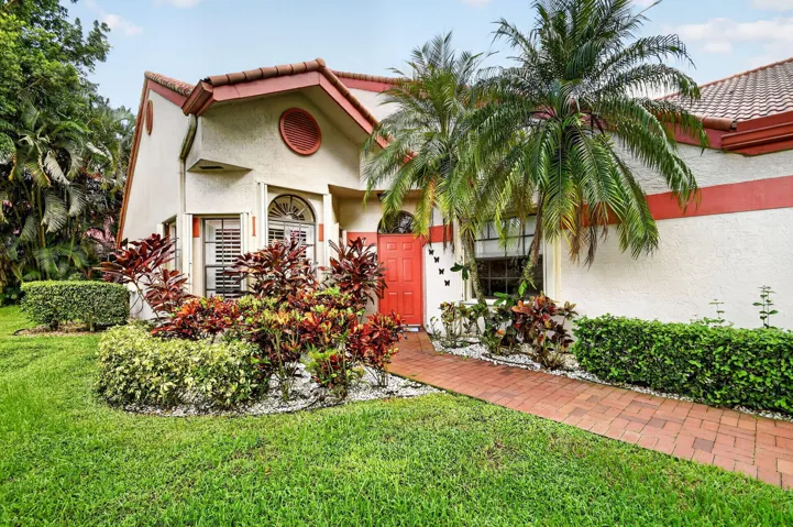 Stucco exterior with a tile roof, featuring a red entry door, a brick paver pathway, and mature landscaping