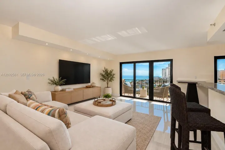 Open-concept living space featuring high-gloss tile flooring, recessed ceiling lighting, a built-in stone-top breakfast bar, and expansive multi-panel sliding glass doors opening to a balcony with an ocean backdrop