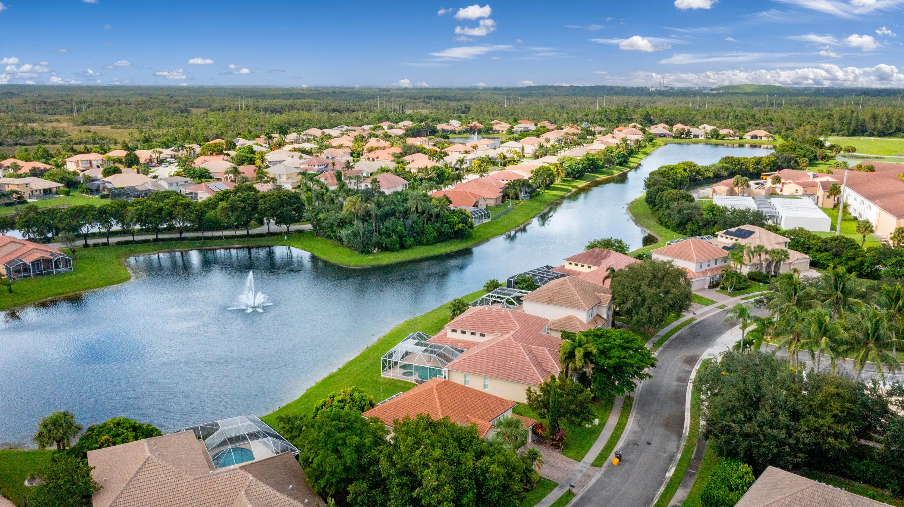 Community featuring a prominent waterway with a fountain, surrounded by residential properties with tiled roofs and screened-in pool enclosures