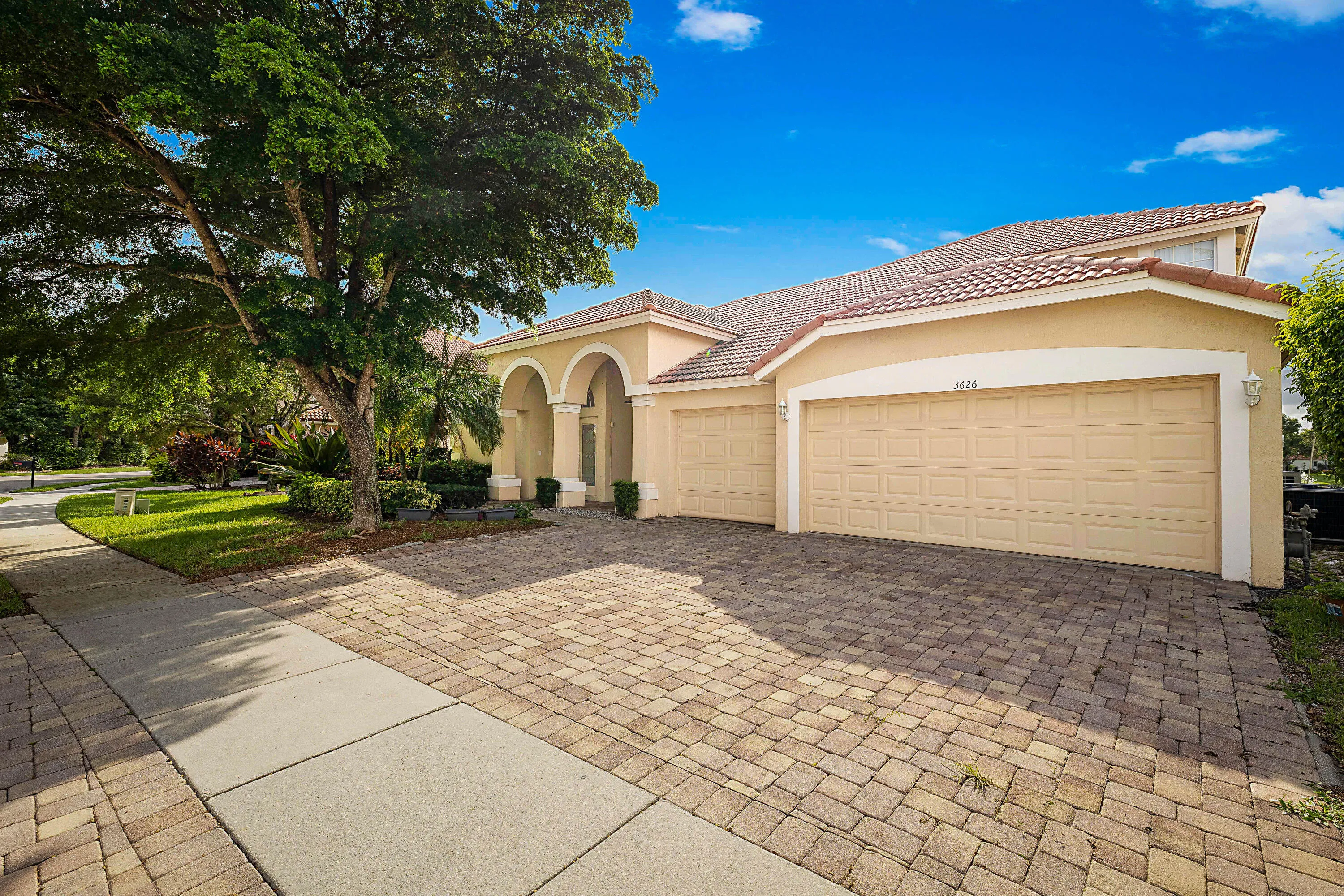 Elegant stucco residence featuring a triple-bay garage with paneled doors, circular driveway with paver hardscaping, and mature landscaping including a large shade tree