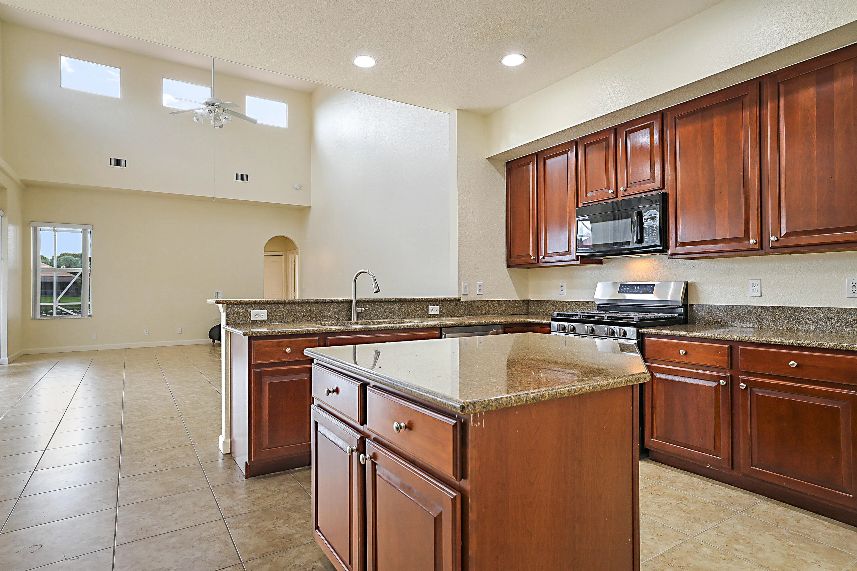 Spacious kitchen featuring rich wood cabinetry, dual islands with granite countertops, stainless steel appliances, recessed lighting, and tile flooring that extends into the adjoining great room with vaulted ceilings and abundant natural light from transom windows