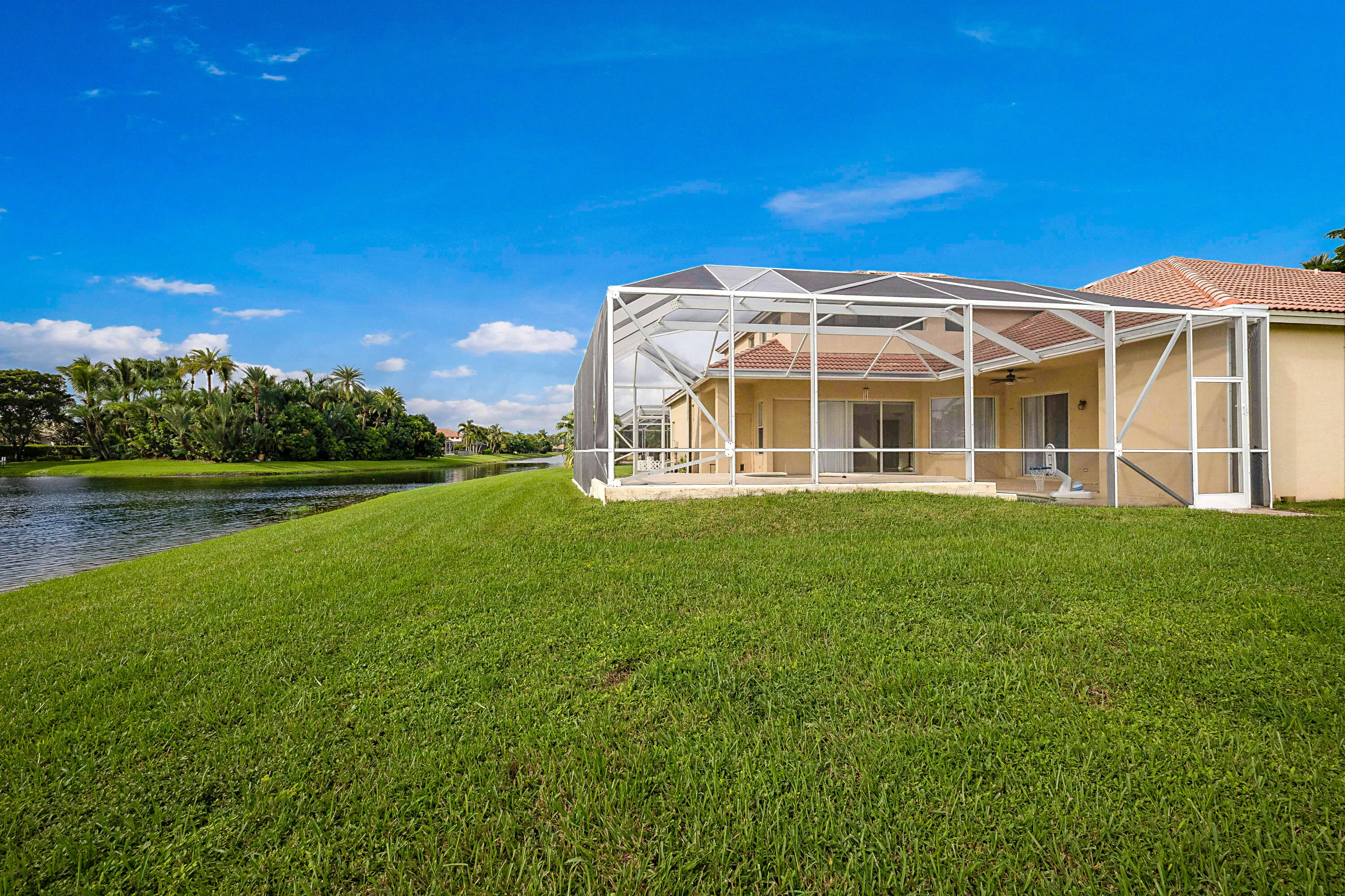 Expansive manicured lawn overlooking a scenic waterfront, complemented by a screened lanai with pitched roof and ceiling fans, attached single-story residence with clay tile roofing, and mature landscaping with native vegetation framing the property