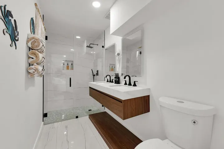 Walk-in shower with clear glass enclosure, a floating wood-finish vanity with dual sinks, and matte black fixtures