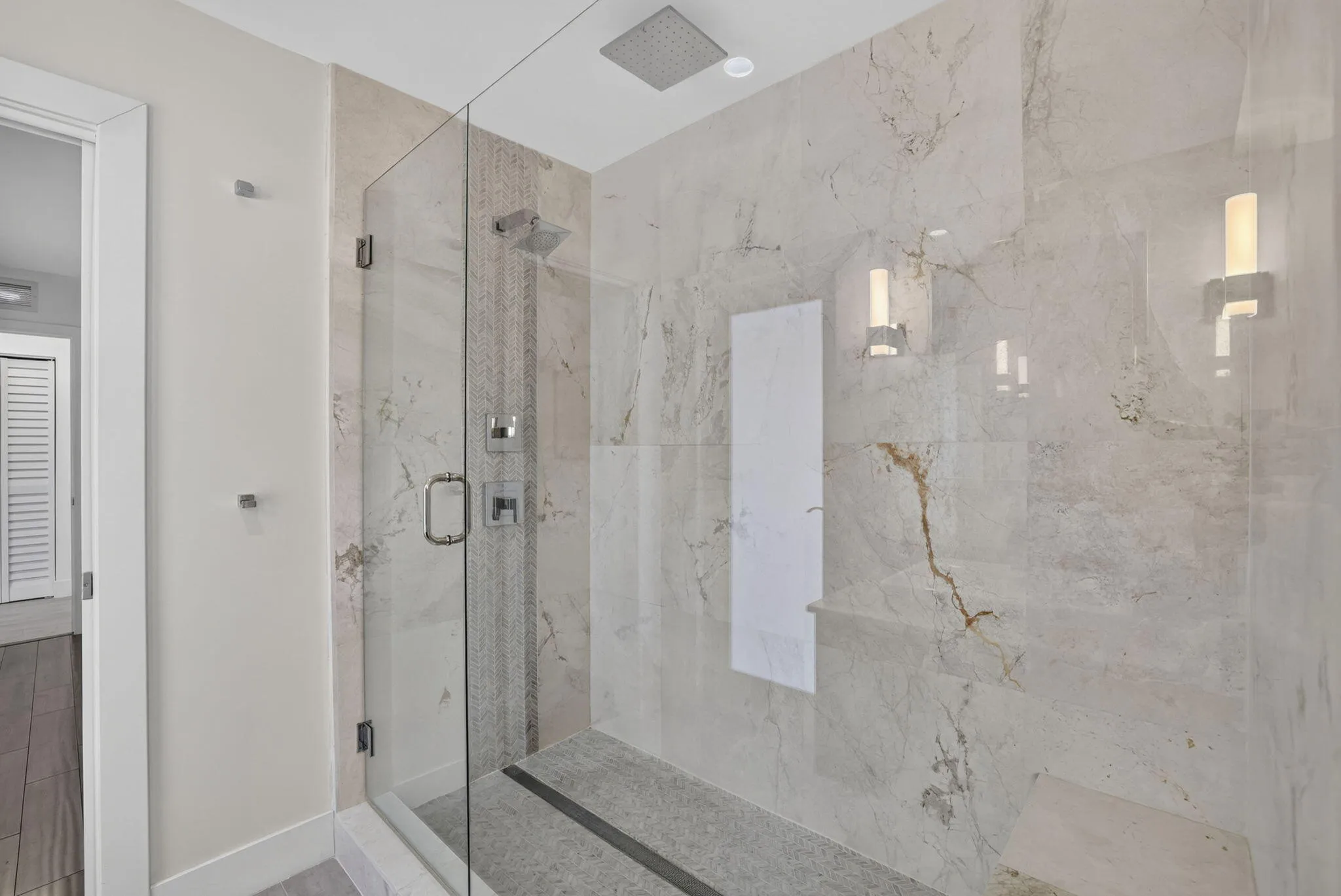 Frameless glass shower enclosure with a built-in bench, ceiling-mounted rain shower head, and wall-mounted sconce lighting