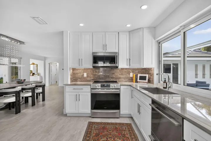 White shaker cabinetry, stainless steel appliances, stone tile backsplash, light-toned flooring, and large window