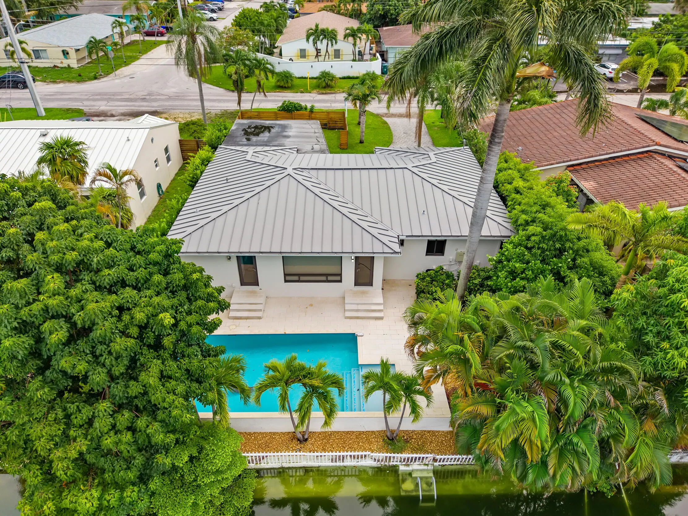 White exterior residence with a metal roof, featuring an in-ground swimming pool, waterside landscaping, and mature trees