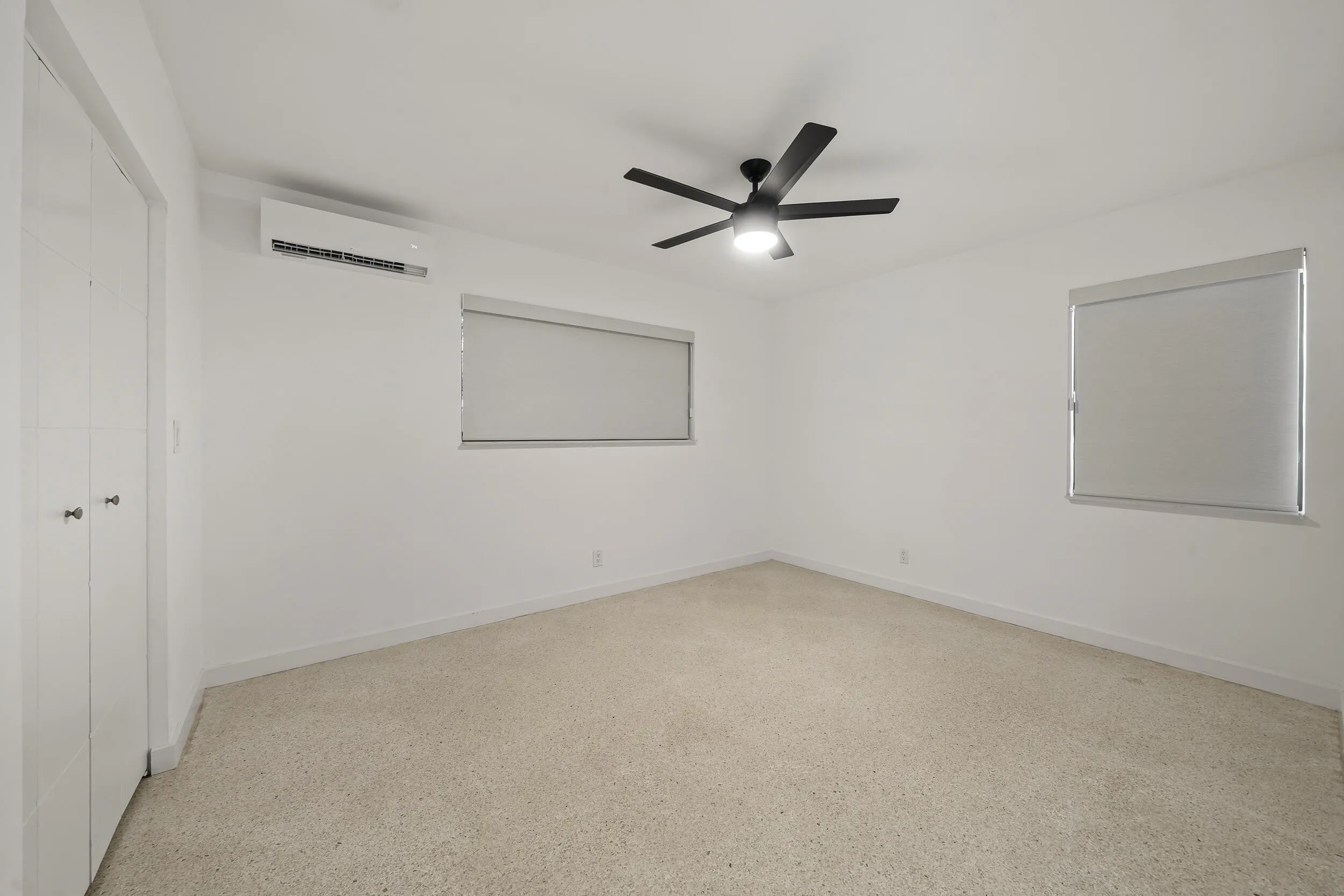 Carpeted room featuring a ceiling fan with integrated lighting, two windows with roller shades, and a wall-mounted air conditioning unit