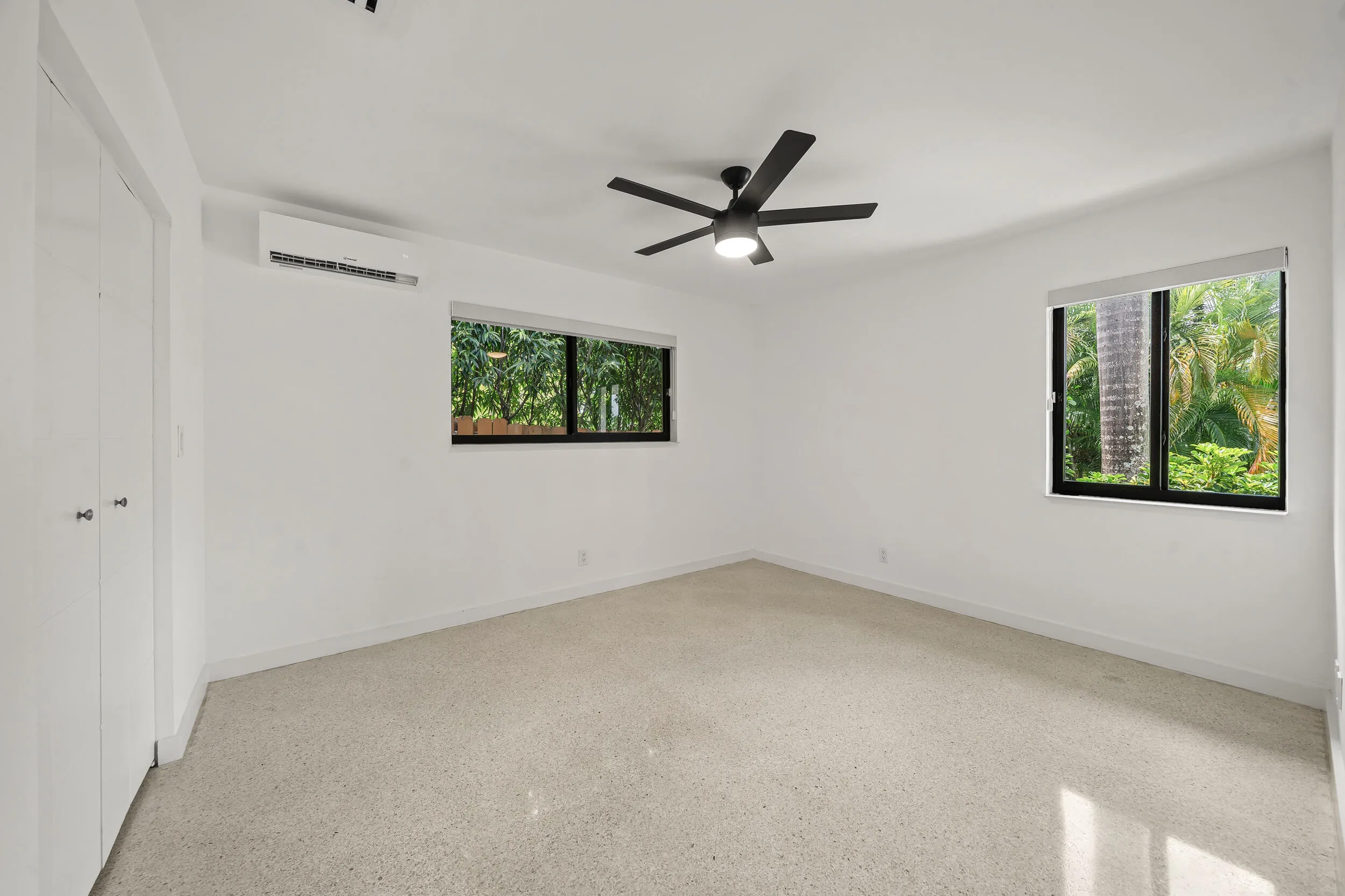 Bright room featuring white walls, terrazzo flooring, a ceiling fan with integrated lighting, a wall-mounted AC unit, and two windows with black frames
