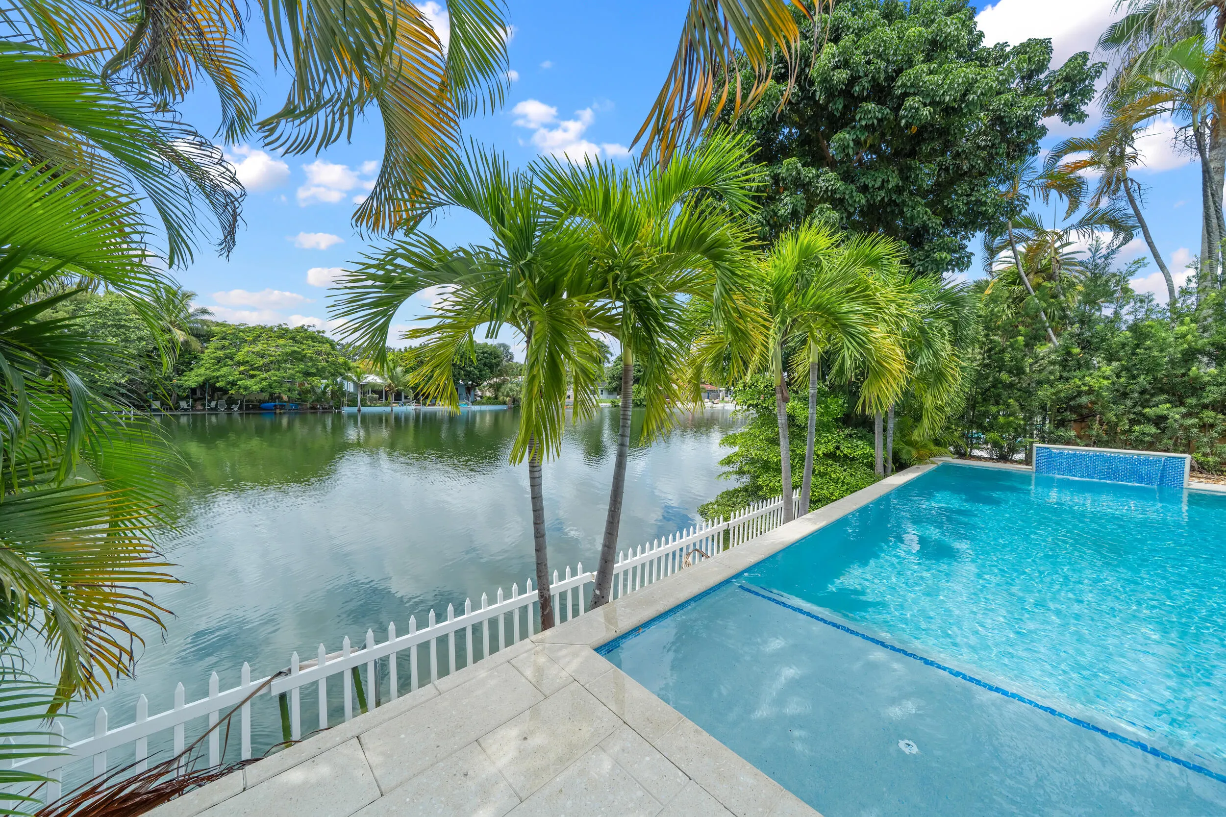 Waterfront property featuring an in-ground swimming pool, white picket fence, and mature palm trees