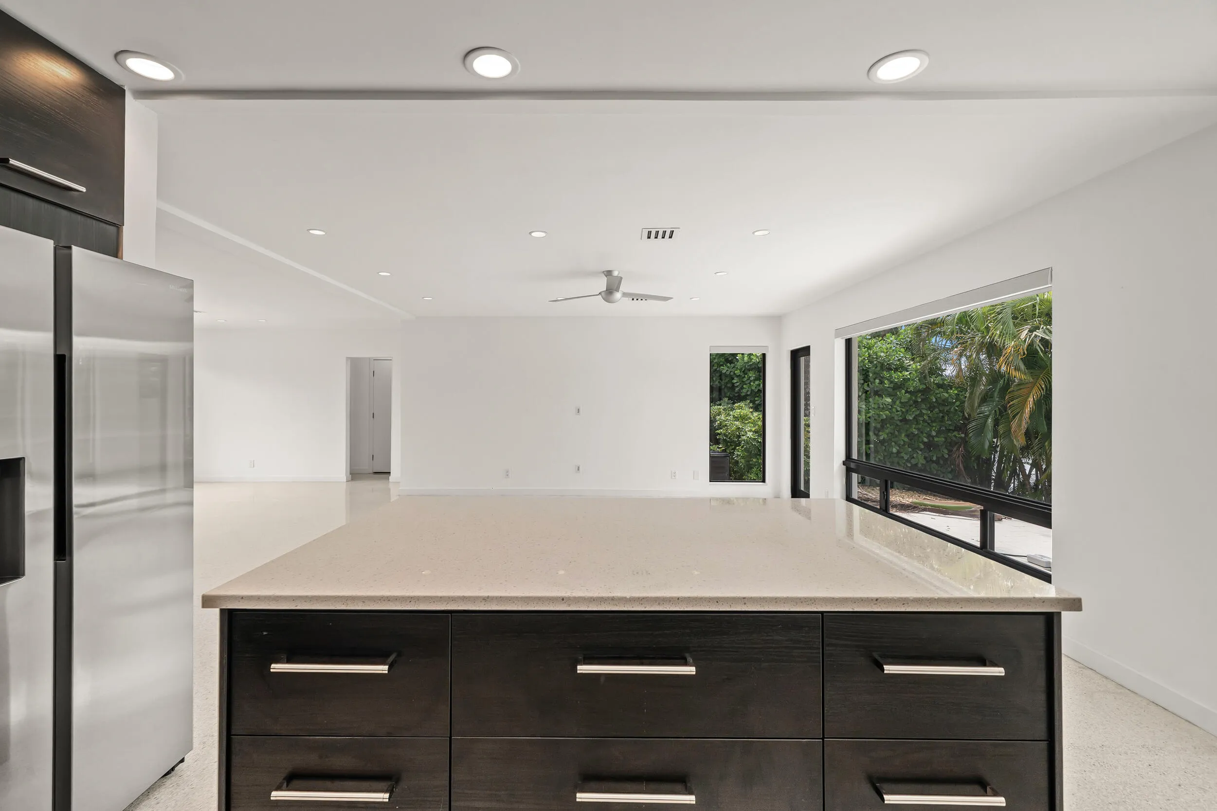 Spacious kitchen island with a light-toned countertop, stainless steel refrigerator, recessed lighting, and large windows with exterior foliage