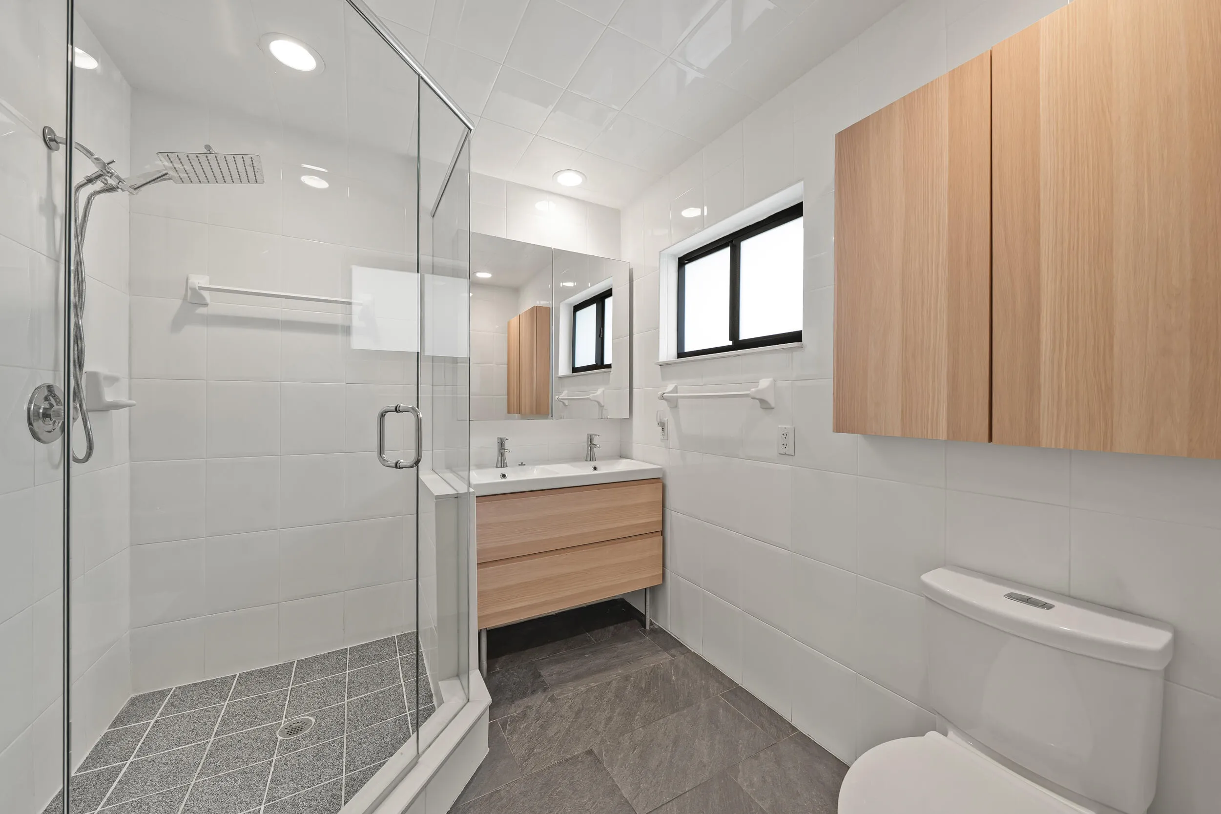 Glass-enclosed shower with rainfall showerhead, dual vanity with undermount sinks, wood-finish cabinetry, and recessed lighting