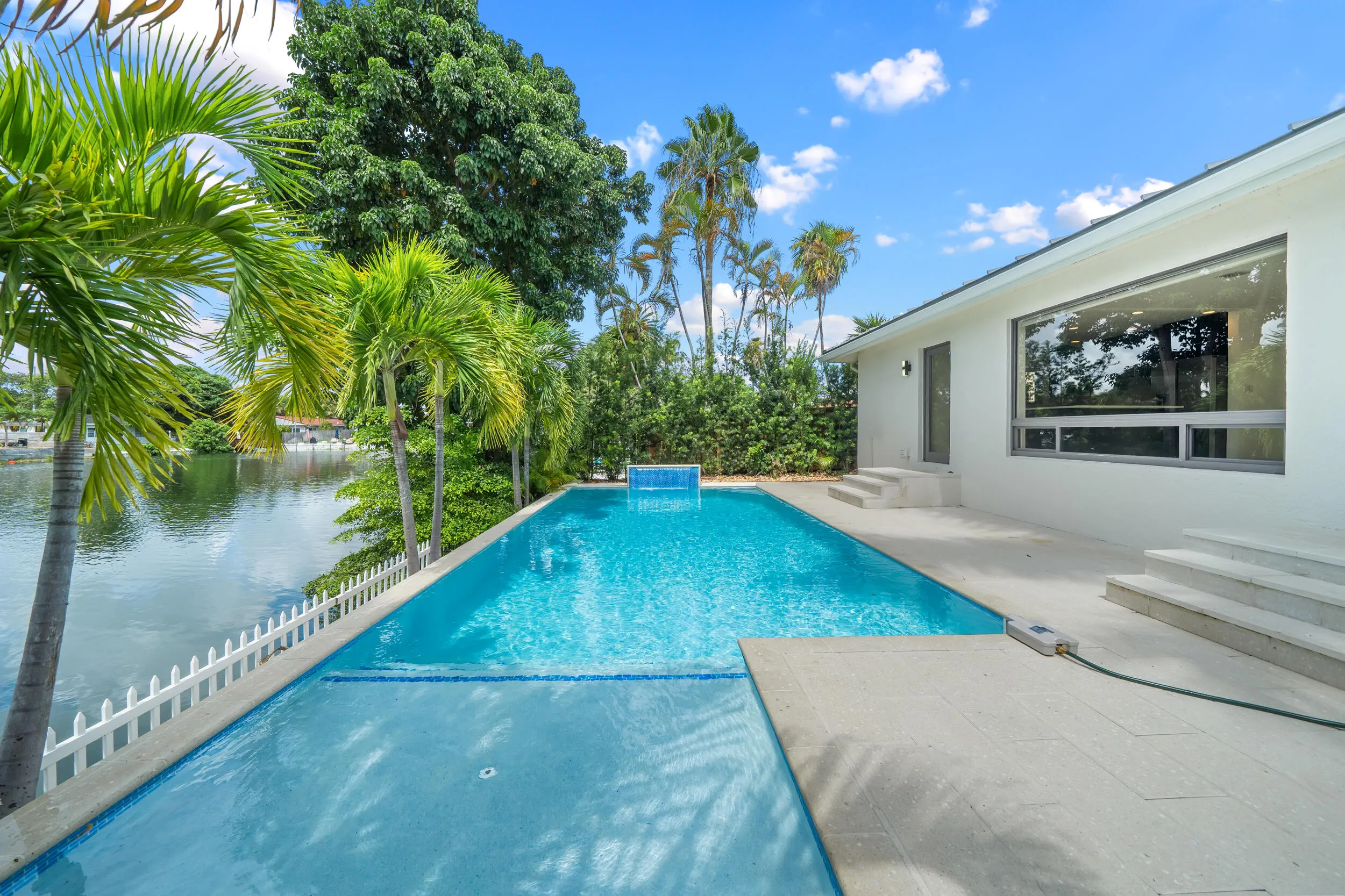 Waterfront property featuring a perimeter fence, mature palm trees, and an in-ground swimming pool with a tile-finish accent