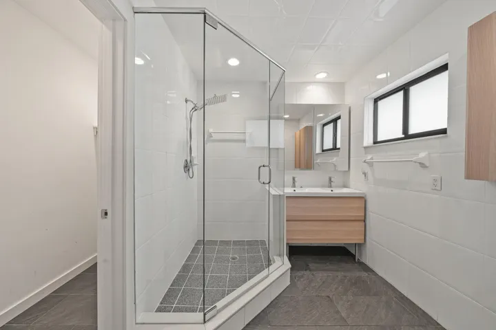 Glass-enclosed shower with a rainfall showerhead, dual sink vanity with integrated storage, full-width mirror, and tiled flooring