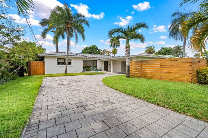 Paved driveway leading to a single-story residence with a white stucco exterior, framed by mature palm trees and a horizontal wood fence