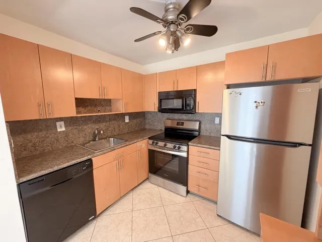 Kitchen featuring light wood-finish cabinetry, stone-look countertops, stainless steel refrigerator, black dishwasher, and a ceiling fan with integrated lighting