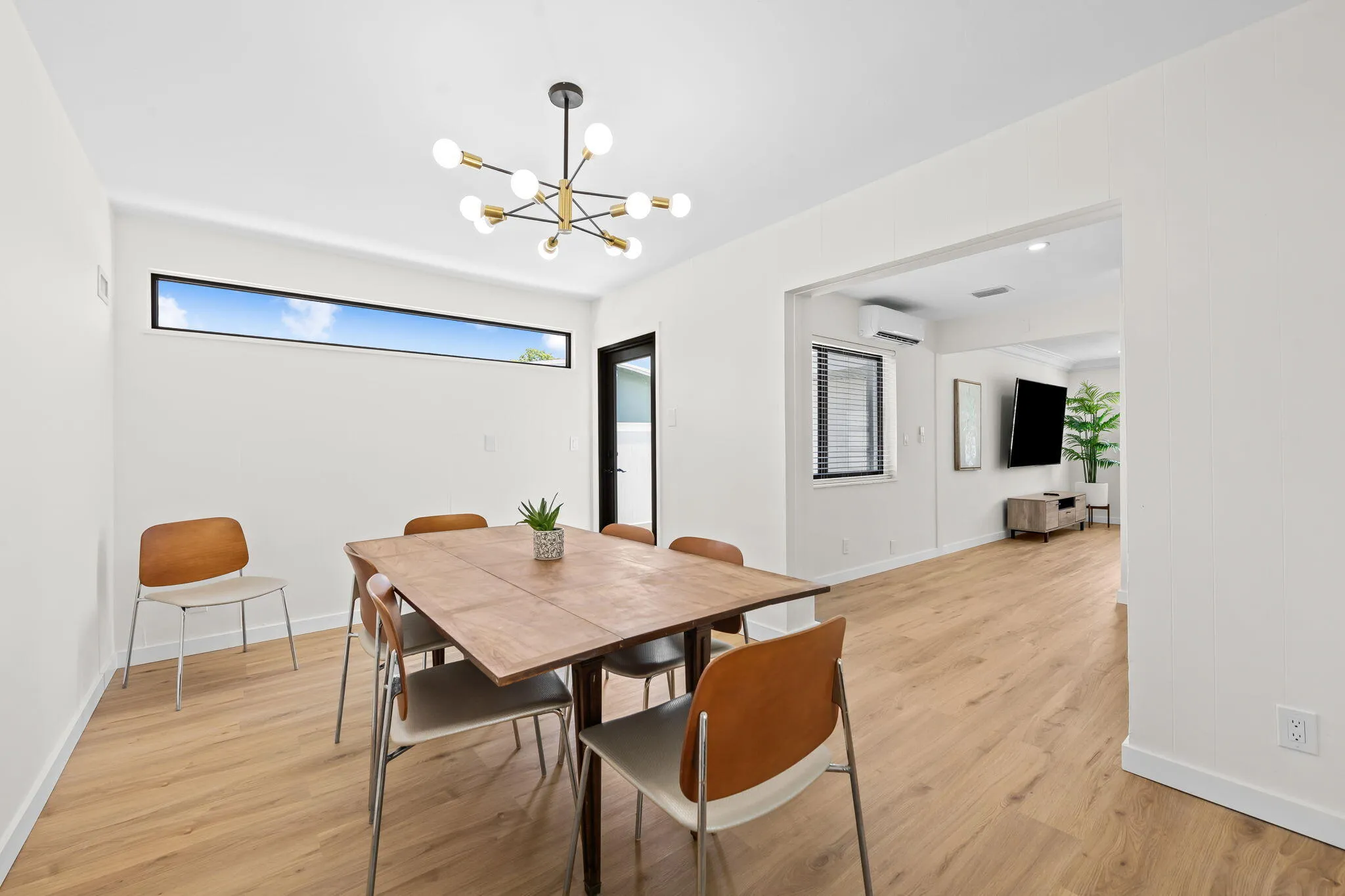 Light wood-finish flooring throughout, modern linear window, contemporary chandelier with multiple light sources, white walls, and a black-framed glass-paneled door