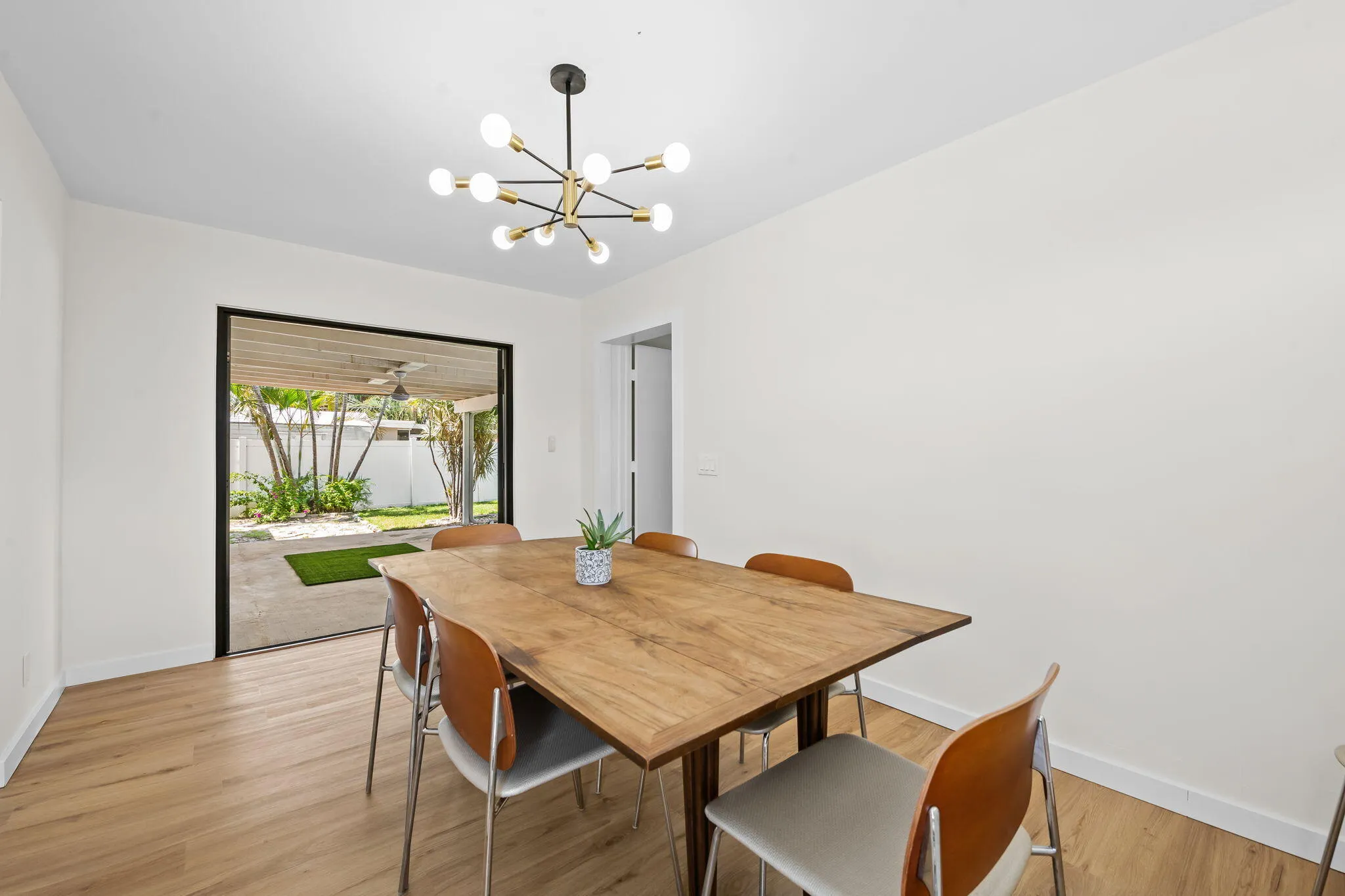 Modern dining space featuring a wood-finish dining table, warm wood flooring throughout, and abundant natural light from large glass doors opening to an outdoor garden area