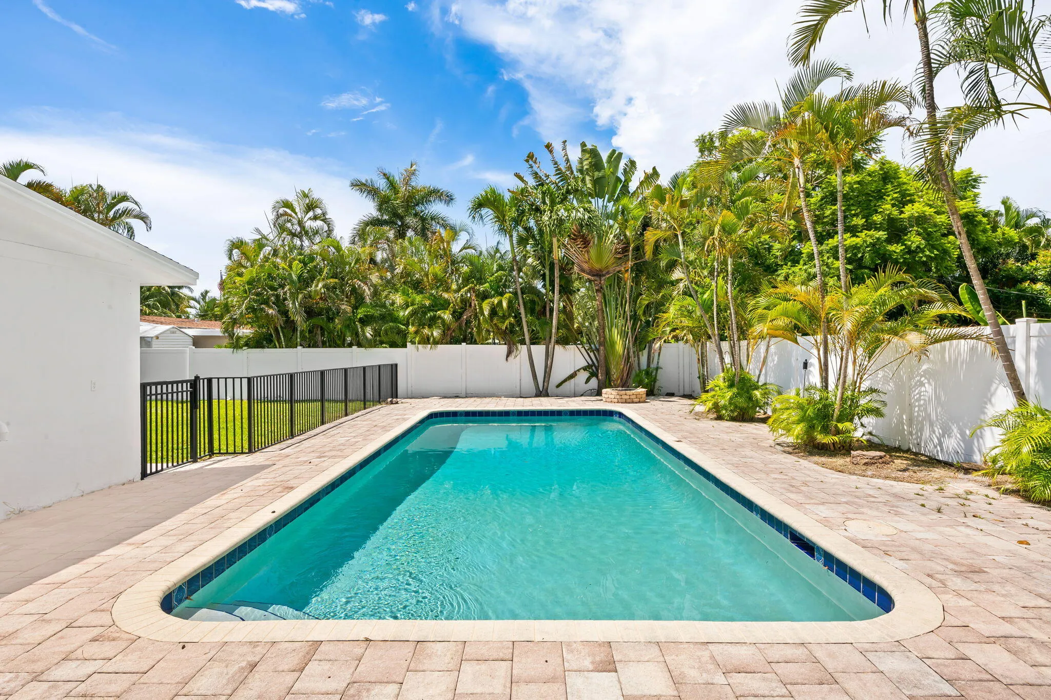 Rectangular swimming pool with curved corners, paver decking, and a black metal fence enclosing a grass area
