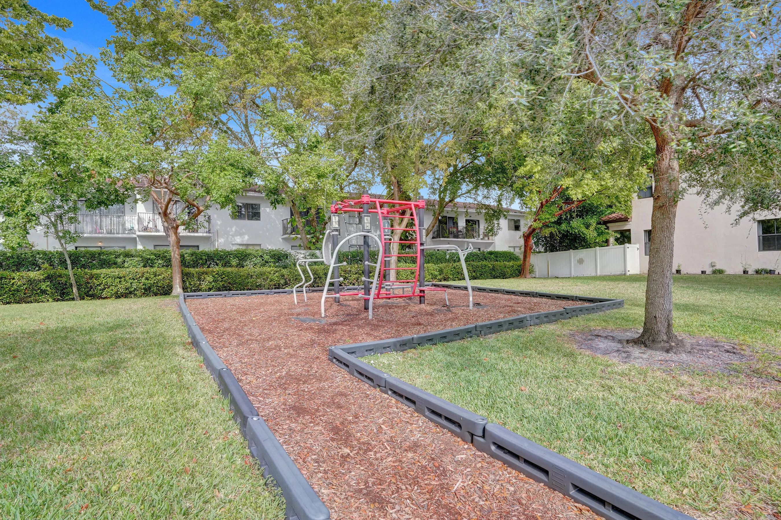 Outdoor recreation area featuring a play structure, wood chip surface, grass, trees, and perimeter curbing