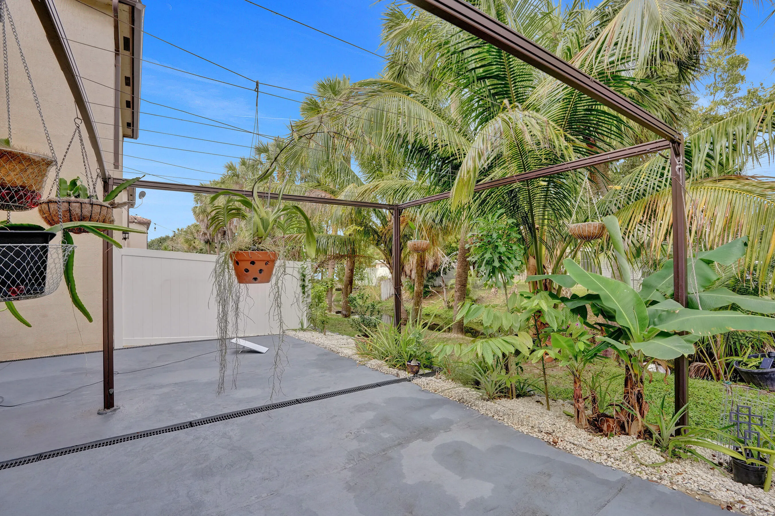 Paved patio area with a pergola structure, bordered by a white privacy fence and lush tropical landscaping
