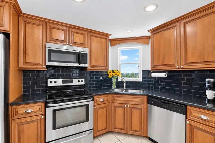 Wood-finish cabinetry with recessed panel doors, stainless steel appliances, dark subway tile backsplash, dark countertops, and a stainless steel sink
