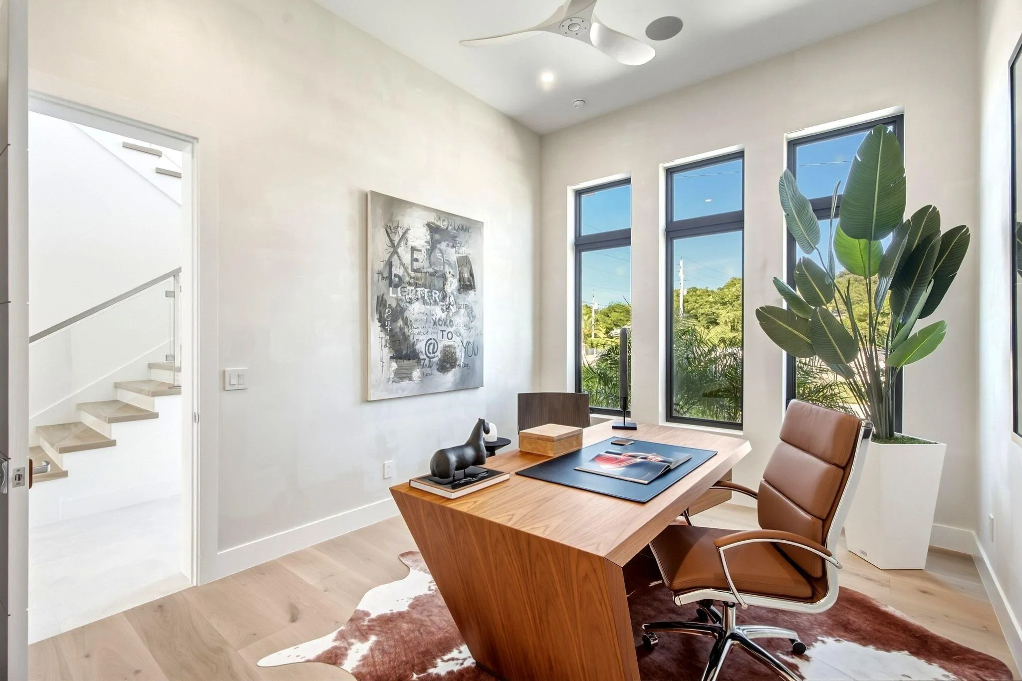 Light wood-finish flooring, three vertical windows with dark frames, modern ceiling fan, recessed lighting, and a floating staircase with a glass railing