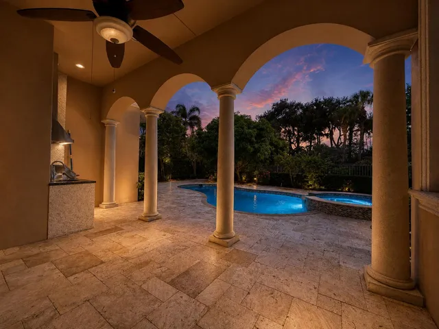 Covered outdoor patio featuring arched openings, stone columns, patterned tile flooring, built-in outdoor kitchen, and ceiling fan