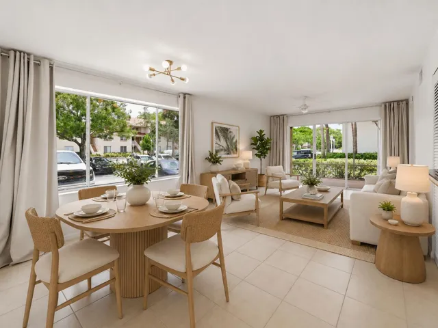 Bright open-concept living and dining room with large windows, natural light, tile flooring, neutral coastal furnishings, and sliding glass doors opening to a ground-floor patio.
